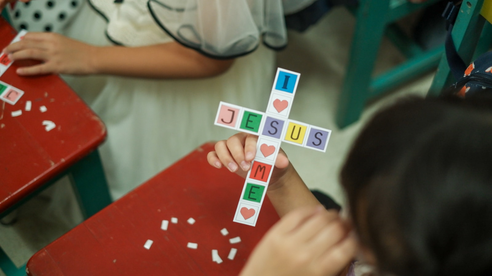 A child holds up a colourful paper cross craft spelling JESUS at a red desk